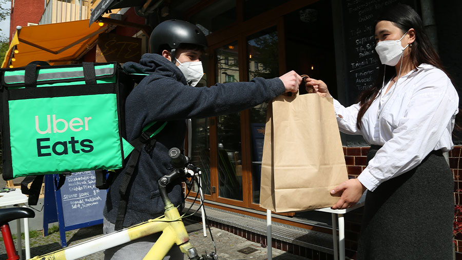Eine junge Frau vor einem Restaurant übergibt einem Fahrradkurier eine Papiertüte. Der Kurier trägt einen türkisfarbenen Rucksack mit der Aufschrift "Uber Eats"