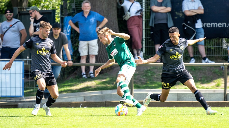 Auf sommerlichem Rasen spielen zwei Fußballer des Bemer SV in dunkelblauer Spielkleidung gegen einen Spieler des VfB Lübeck in weiß-grüner Spielkleidung