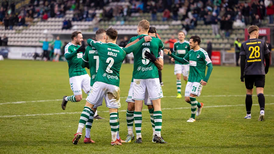 Mehrere Spieler des VfB Lübeck in weiß-grüner Spielkleidung umarmen sich auf einem Fußballplatz. Ein Spieler in dunkelblauer Spielkleidung ist am Rand zu sehen