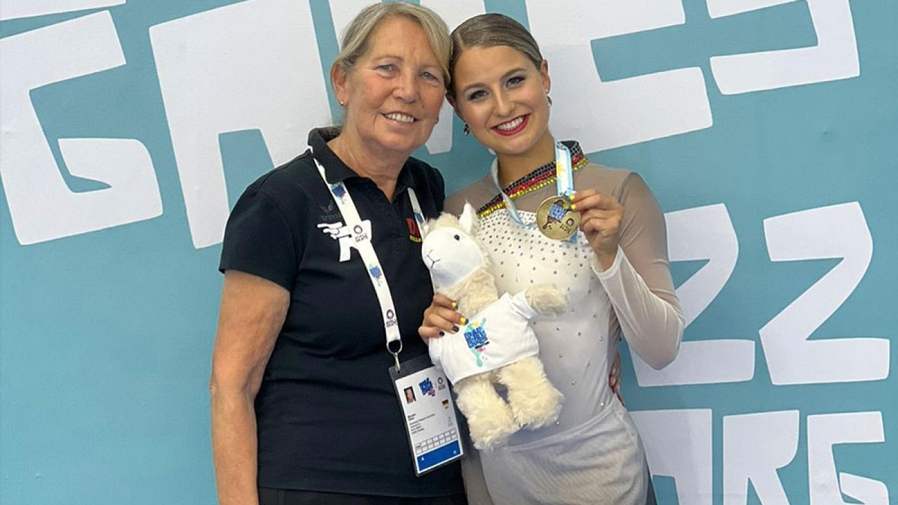 Eine junge Frau in Turndress zeigt stolz eine goldene Medaille. Eine ältere Frau mit Akkreditierung um den Hals hält sie im Arm