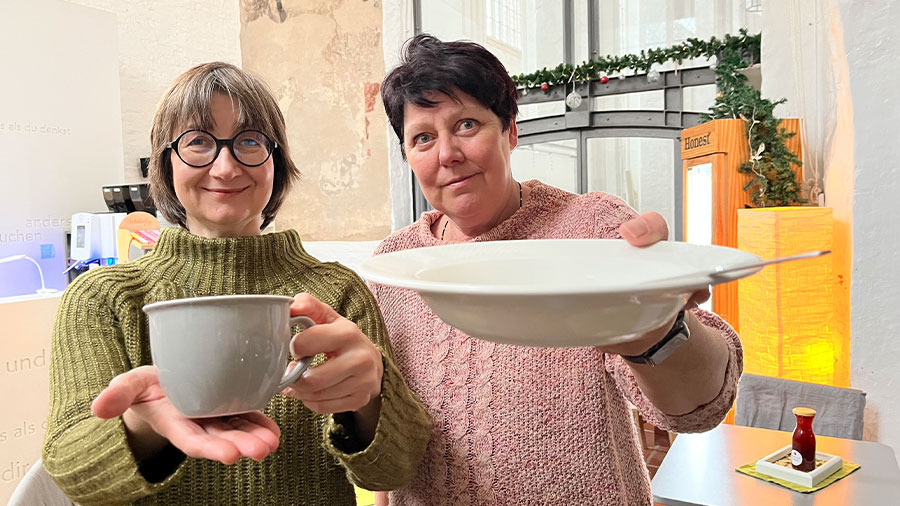 Zwei Frauen in Strickpullovern halten eine graue Tasse und einen Teller mit Löffel in die Kamera