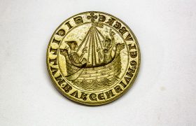 Ein goldenes Stadtsiegel der Hansestadt Lübeck mit einem Boot und zwei Personen drauf.