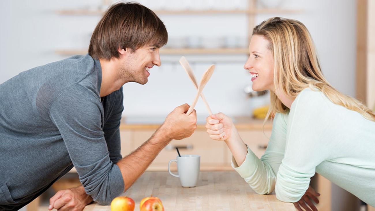 Ein Mann und eine Frau lehnen auf einer Tischplatte gegenüber. Sie lächeln sich an und kreuzen hölzerne Kochlöffel