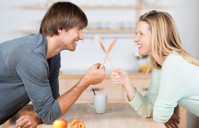 Ein Mann und eine Frau lehnen auf einer Tischplatte gegenüber. Sie lächeln sich an und kreuzen hölzerne Kochlöffel
