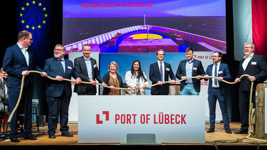 Mehrere Menschen auf einer Bühne halten gemeinsam ein Tau fest. Im Vordergrund steht auf einem Schild "Port of Lübeck"