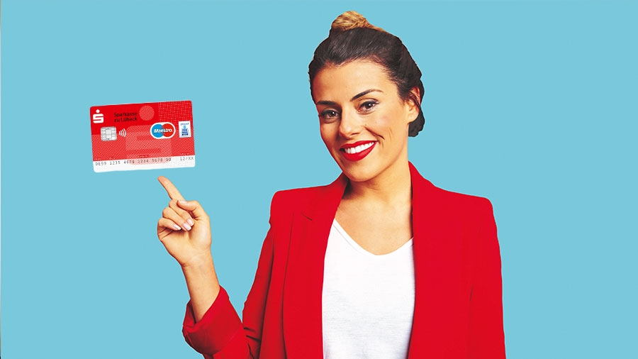 Eine Frau mit rotem Jacket zeigt auf eine Girokarte der Sparkasse, die in der Luft schwebt, dabei schaut sie in die Kamera.