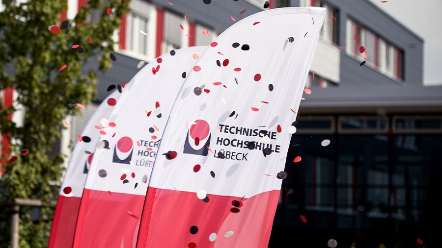Vor einem Bürogebäude stehen drei weiß-rote Beachflags mit der Aufschrift "Technische Hochschule Lübeck". Davor fliegt Konfetti durch die Luft.