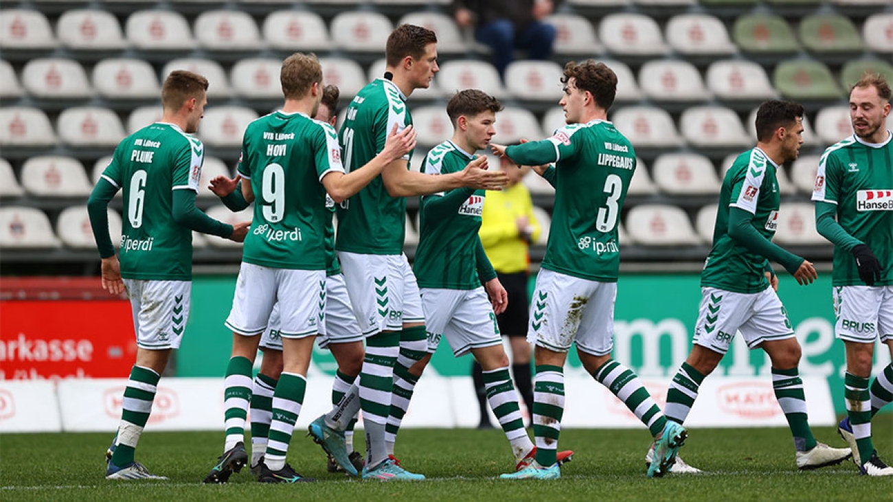 Mehrere Spieler des VfB Lübeck in grünen Trikots mit weißen Hosen beglückwünschen sich gegenseitig auf dem Fußballplatz. Im Hintergrund sieht man mehrere leere Zuschauerreihen