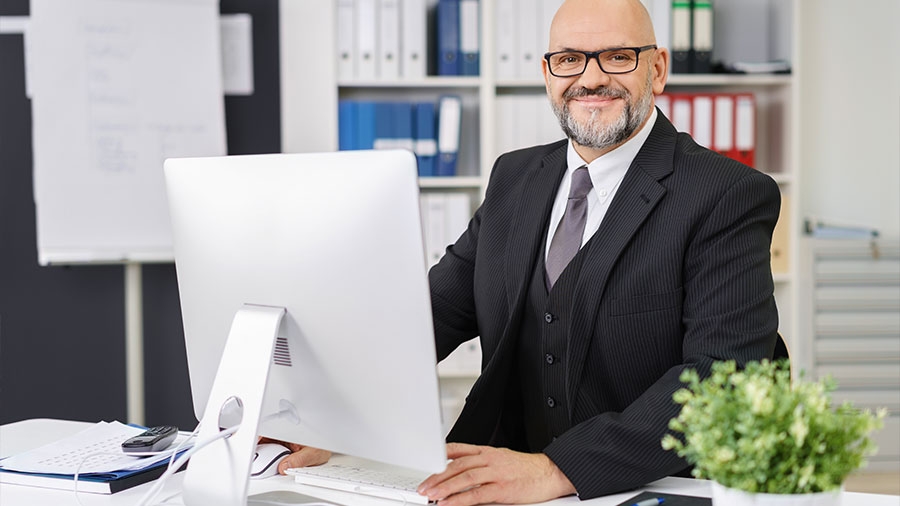Ein Mann mit grau meliertem Vollbart und schwarzer Brille sitzt im eleganten Dreiteiler in einem Büro hinter einem Computer und lächelt freundlich in die Kamera