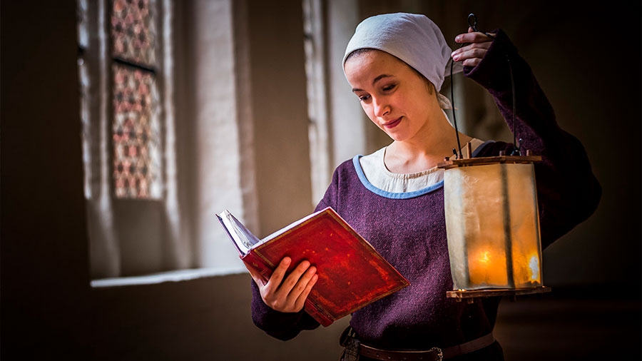 Eine Frau mit mittelalterlicher Kleidung hält ein geöffnetes Buch in der Hand und eine Laterne.