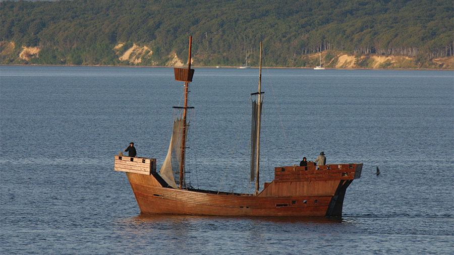 Eine nachgebaute Kogge fährt in der Nähe der Küste mit drei Personen an Bord.