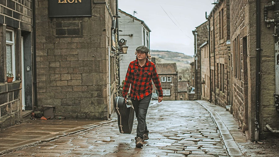Ein Mann mit rotem Hemd und einer Gitarre läuft eine alte englische Straße hinauf.