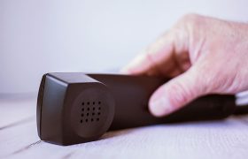 Eine Hand einer alten Person legt den Hörer eines Telefons auf eine Holzfläche.