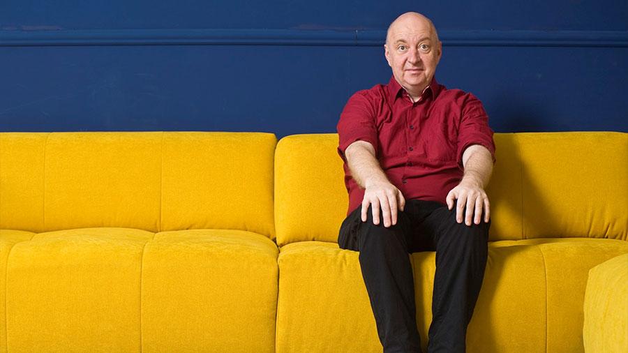 Ein Mann mit rotem Hemd sitzt auf einem gelben Sofa vor einer blauen Wand.