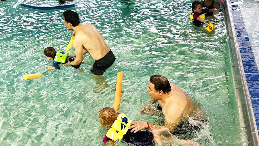 Mehere Väter üben mit ihren Kindern schwimmen in einem flachen Becken.