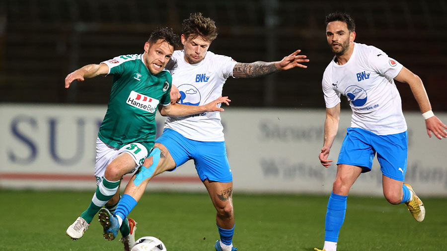 Ein Spieler des VfB Lübeck und zwei von Blau-Weiß Lohne laufen dem Ball hinterher.