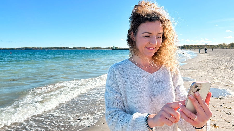 Eine Frau steht am Strand und bedient ihr Handy, während der Wind durch ihre lockigen Haare weht.