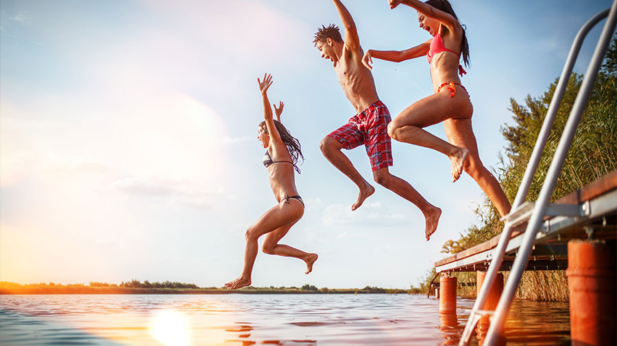 Drei junge Menschen springen von einem Steg ins Wasser, die Sonne scheint.