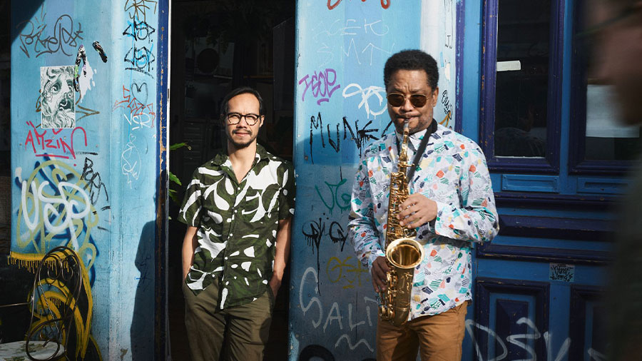 Zwei Männer vor einem blauen Gebäude. Einer spielt Saxophone.