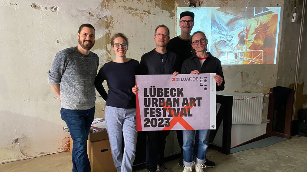 5 Personen stehen in einem Gemäuer an dessen Wand ein Bild gestrahlt wird. Sie halten ein Plakat für das Urban Art Festival.