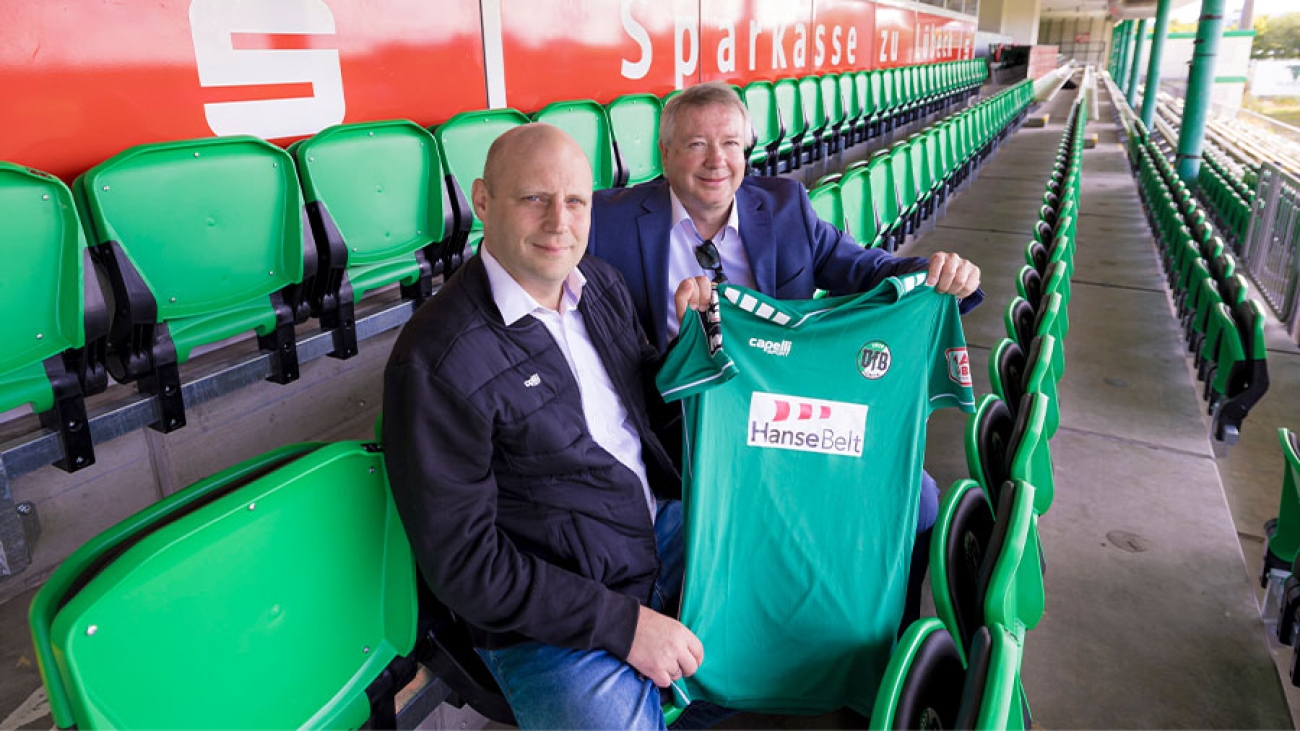 Zwei Männer halten ein Trikot auf der Sparkassen Tribüne.